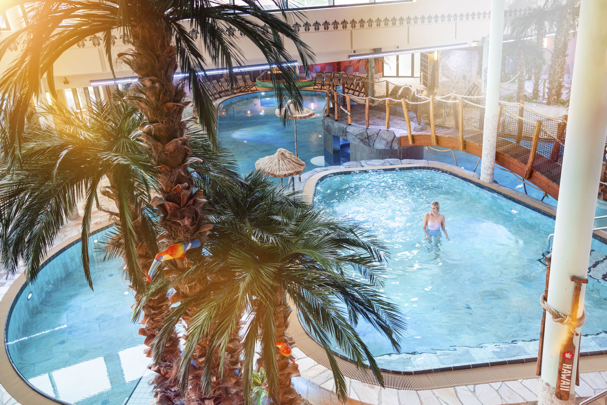 Tropisch gestaltetes Freizeitbad mit Innenpool, Palmen und Holzbrücke; eine Frau steht im Wasser.