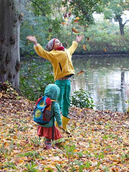 Zwei Kinder spielen im Herbstlaub am Ufer eines Sees. Eines wirft Blätter in die Luft, während das andere zuschaut. Bunte Kleidung und herbstliche Stimmung.