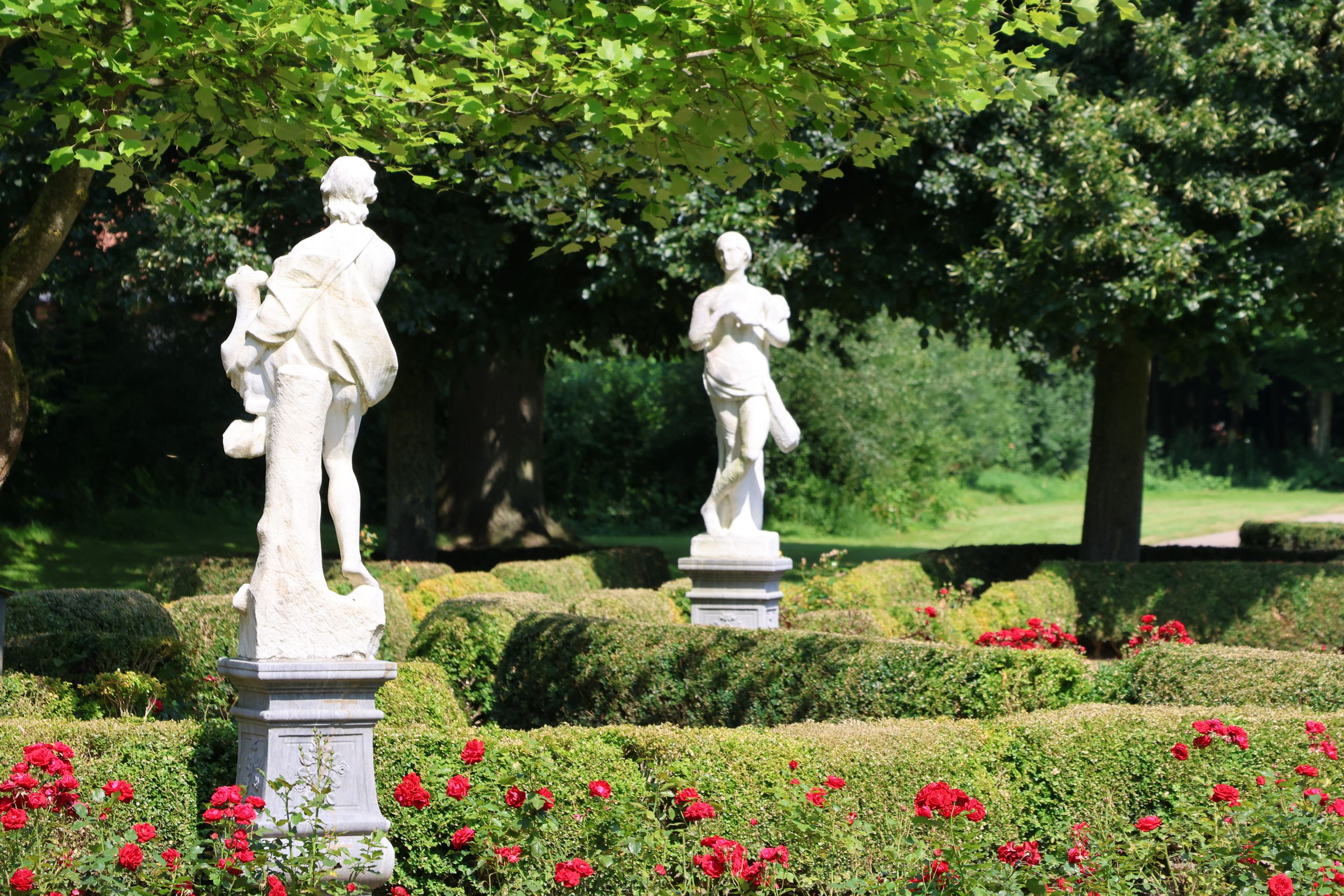 Zwei weiße Statuen stehen in einem gepflegten Garten, umgeben von roten Rosen und grünen Hecken. Im Hintergrund sind Bäume zu sehen.