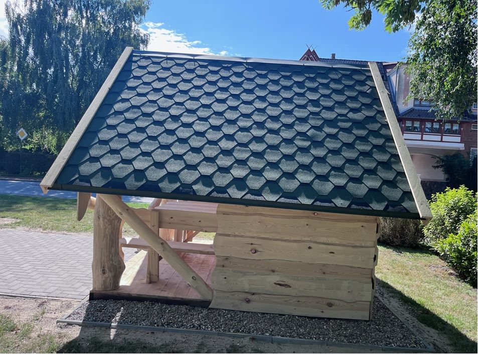 Schutzhütte neben einem Gehweg, im Hintergrund Bäume und ein Wohnhaus.