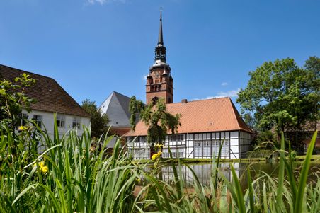 Historisches Gebäude mit Fachwerk und Kirchturm, umgeben von grüner Vegetation und einem Teich in Itzehoe.