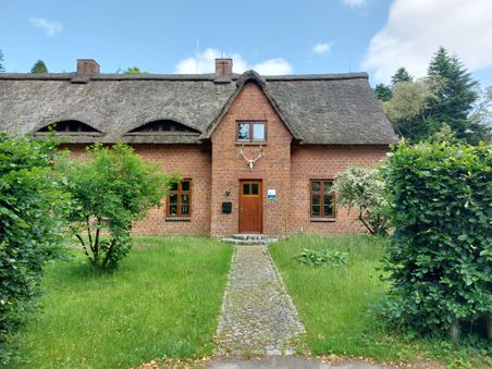 Ein Backsteinhaus mit Reetdach und gepflegtem Gartenweg, umgeben von grünen Bäumen und Sträuchern. Der Himmel ist leicht bewölkt.