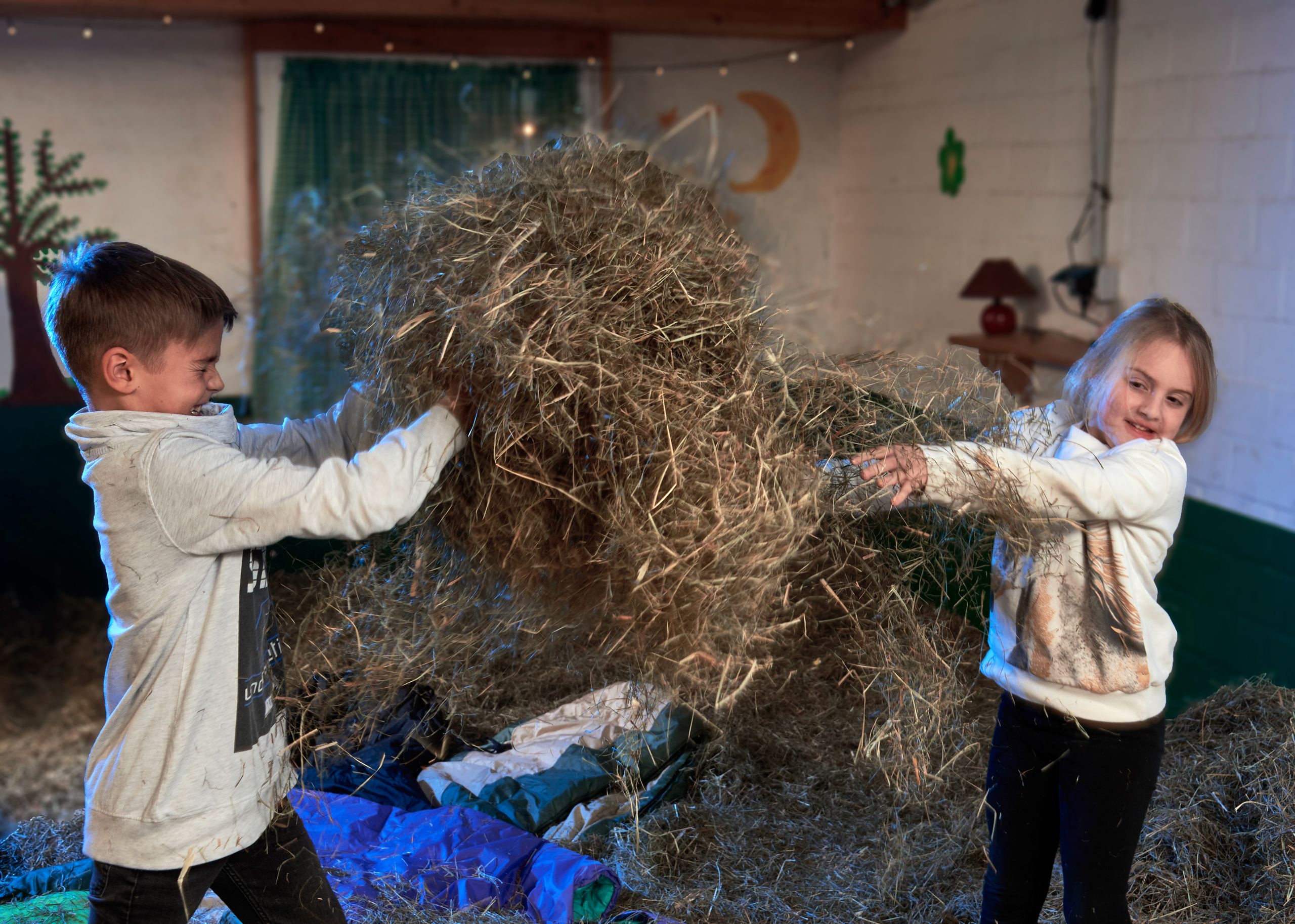 Zwei Kinder spielen im Stall mit Heu und werfen es in die Luft. Im Hintergrund sind Lichter, eine grüne Gardine und Wandmalereien zu sehen.