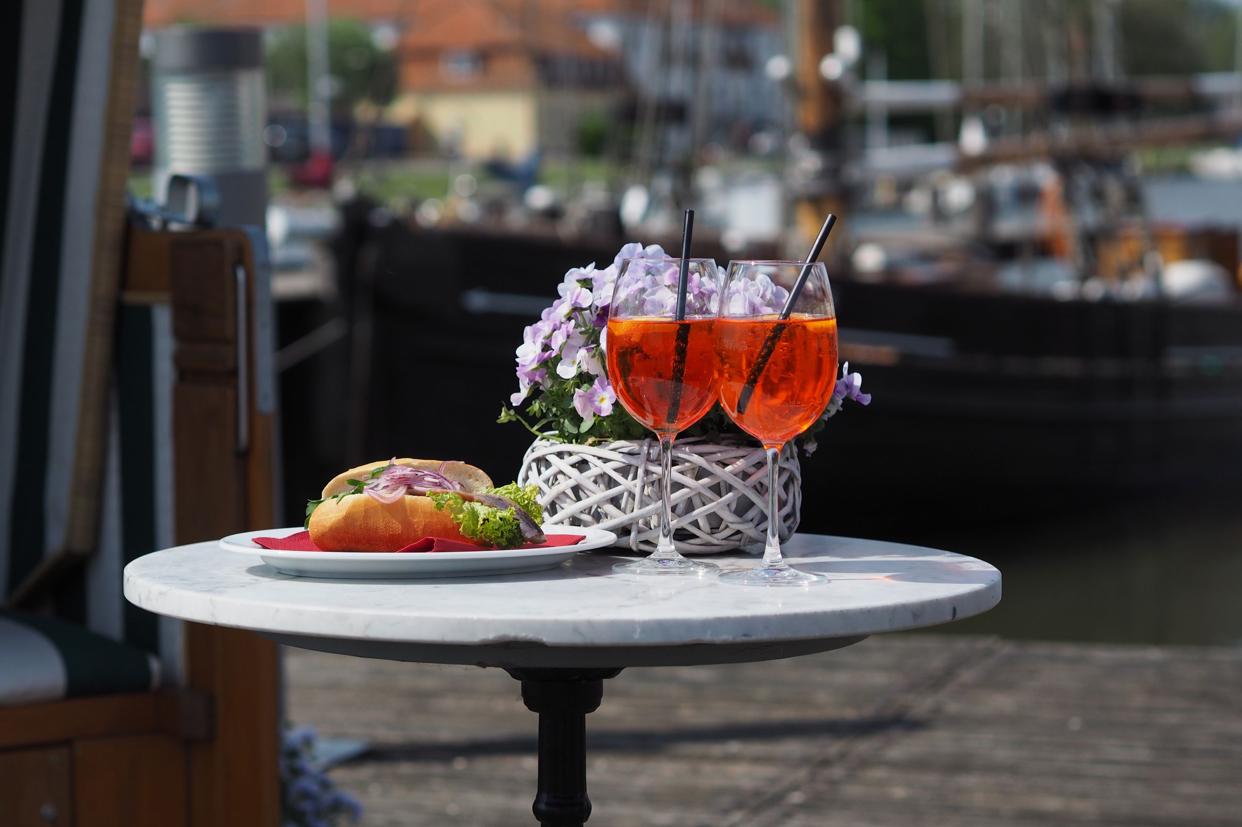 Ein Matjesbrötchen und zwei Aperol Spritz auf einem Tisch am Hafen, dekoriert mit einem Blumenkorb.