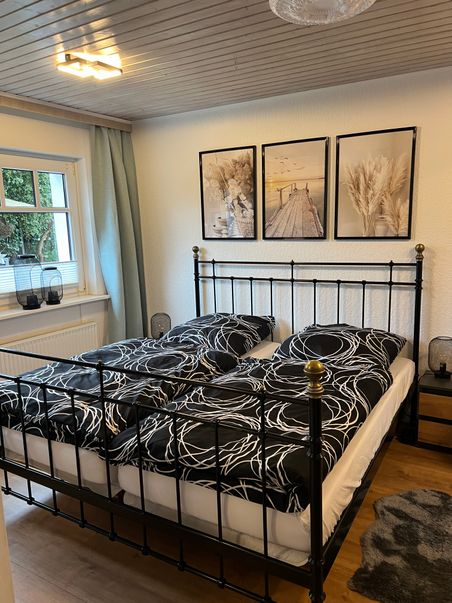 Gemütliches Schlafzimmer mit schwarzem Metallbett, schwarz-weißer Bettwäsche und drei gerahmten Naturmotiven über dem Bett.