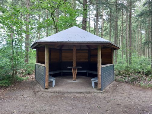 Offener Holzpavillon im Wald mit umlaufenden Bänken und zentralem Tisch unter einem hellen Dach.