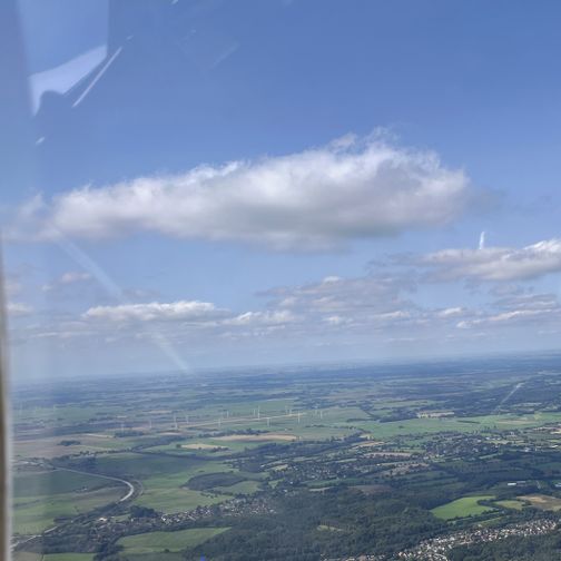 Luftaufnahme einer ländlichen Landschaft mit Feldern, Windrädern und kleinen Ortschaften unter blauem Himmel mit Wolken.