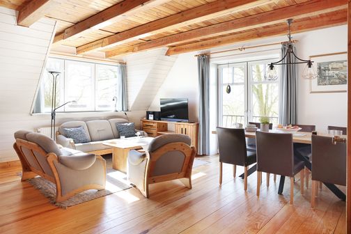 Heller Wohn- und Essbereich mit Holzbalkendecke, Sofa, Sesselgruppe, Fernseher und Esstisch am Fenster.