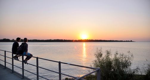 Zwei Personen sitzen auf einem Geländer und genießen den Sonnenuntergang über der Elbe. Der Himmel ist in warmen Farben getaucht, das Wasser spiegelt das Licht.