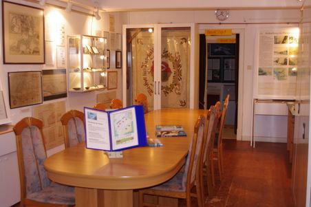 Ausstellungsraum „Dörpstuuv 1“ mit Holztisch, Stühlen, Vitrinen, Karten und historischen Tafeln.