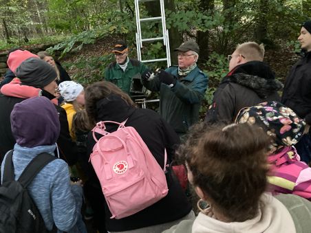 Gruppe bei einer Waldführung: Zwei ältere Männer erklären etwas vor einer Leiter, umringt von Kindern und Erwachsenen im Wald.