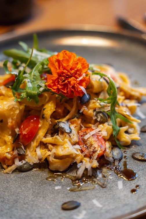 Ein kunstvoll angerichtetes Pastagericht mit einer orangefarbenen Blüte, Rucola, Kürbiskernen und Kirschtomaten auf einem grauen Teller.