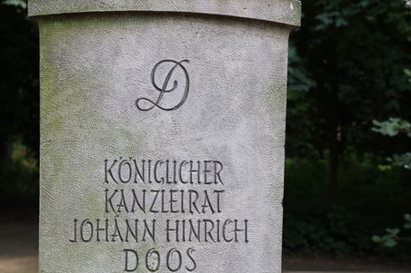 Ein Grabstein mit der Inschrift 'Königlicher Kanzleirat Johann Hinrich Doos' steht in einem grünen, bewaldeten Bereich.