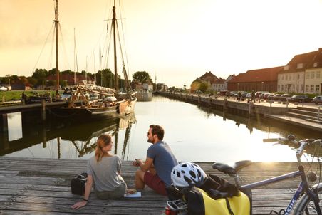 Ein Paar genießt den Sonnenuntergang am Hafen von Glückstadt. Fahrräder und ein Segelboot im Hintergrund schaffen eine entspannte Atmosphäre.