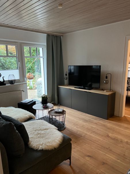Gemütliches Wohnzimmer mit Sofa, Couchtischen, TV-Schrank und großen Fenstern mit Blick in den Garten.