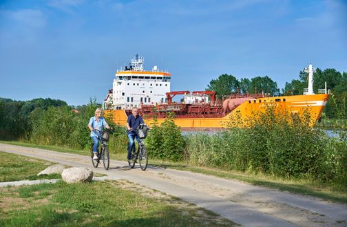 Ein Paar ist mit dem Fahrrad am Nord-Ostsee-Kanal unterwegs. Im Hintergrund ist ein großer oranger Tanker zu sehen.