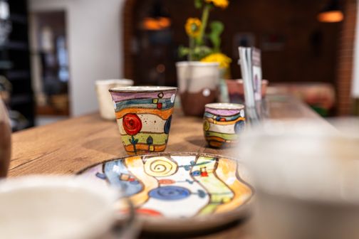 Bunt bemalte Keramikbecher und Teller auf einem Holztisch, im Hintergrund unscharf Blumen und Innenraum.