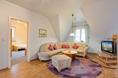 Gemütliches Wohnzimmer mit floralem Ecksofa, rundem Holztisch und Fernseher. Ein Schlafzimmer ist durch eine offene Tür sichtbar.