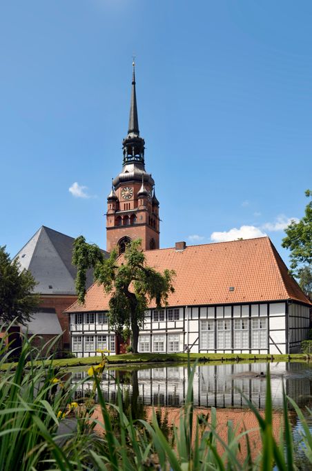 Fachwerkgebäude und Kirchturm in Itzehoe, umgeben von einem Teich und grüner Vegetation. Der klare Himmel betont die Architektur.
