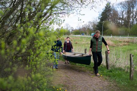 Kanuvorbereitung: Drei Personen tragen ein grünes Kanu auf einem Feldweg durch eine ländliche Landschaft.
