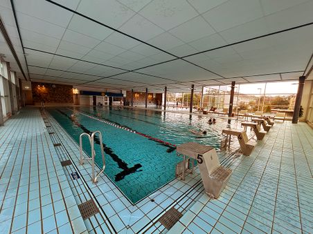 Ein Hallenbad mit einem großen Schwimmbecken, Startblöcken und einigen Schwimmern im Wasser. Große Fenster lassen Tageslicht herein.