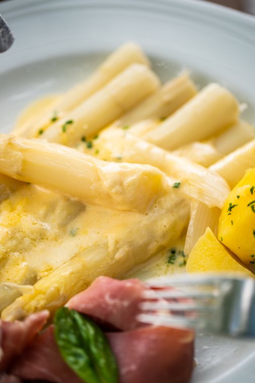 Spargel mit Sauce Hollandaise, serviert mit Kartoffeln und Schinken. Ein klassisches Gericht, das auf einem weißen Teller angerichtet ist.