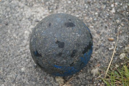 Schwarze Bossel-Kugel mit blauen Farbresten liegt auf grobem Asphalt.