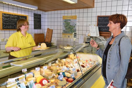 In einem Käseladen reicht eine Verkäuferin einer Kundin ein Stück Käse über der Theke. Im Hintergrund stehen Tafeln mit Sorten und eine Karte der Käsestraße Schleswig-Holstein.