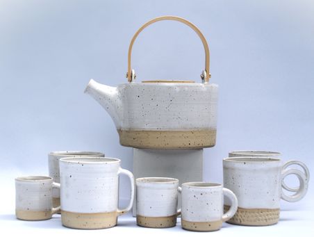 Weiße Keramik-Teekanne mit Holzgriff, umgeben von passenden Tassen. Schlichtes, modernes Design mit natürlichen Farbtönen.