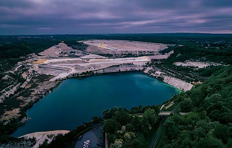 Luftaufnahme eines Steinbruchs mit einem großen, blauen See, umgeben von grüner Landschaft und Hügeln unter bewölktem Himmel.