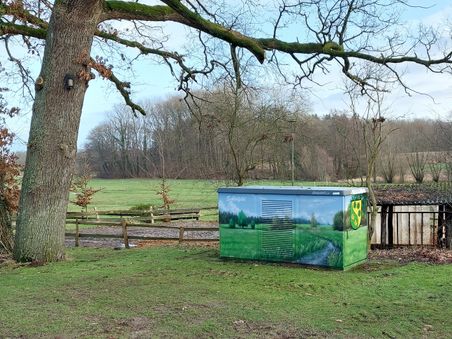 Ein bemalter Stromkasten mit Landschaftsmotiv und Wappen steht neben einem großen Baum auf einer Wiese. Im Hintergrund sind Bäume und ein Zaun zu sehen.