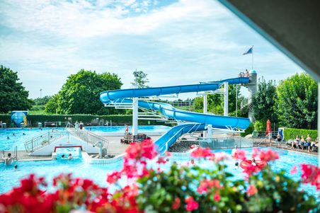 Ein Freibad mit einer großen blauen Wasserrutsche, umgeben von Bäumen. Menschen schwimmen im Wasser. Rote Blumen im Vordergrund.