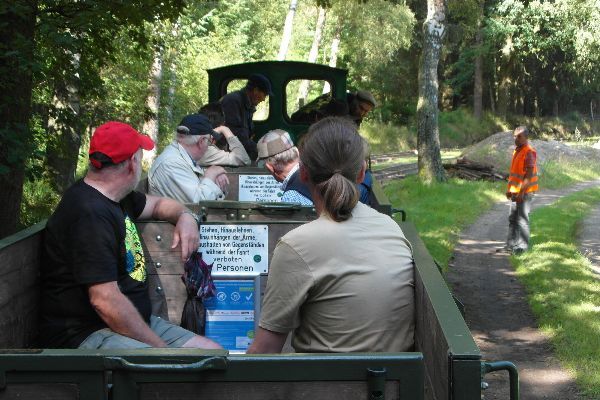 Fahrgäste sitzen in der Moorbahn auf Tour durch den Wald; ein Mann in orangefarbener Warnkleidung steht am Weg.