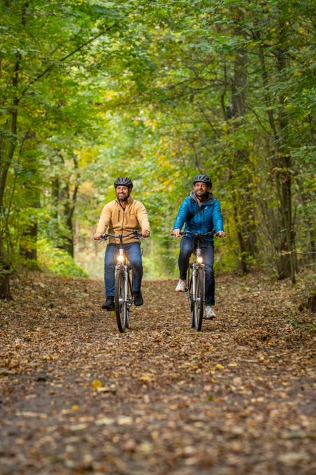 Zwei Männer fahren mit Fahrrädern auf einem mit Laub bedeckten Waldweg. Sie tragen Helme und sind von grünen Bäumen umgeben.