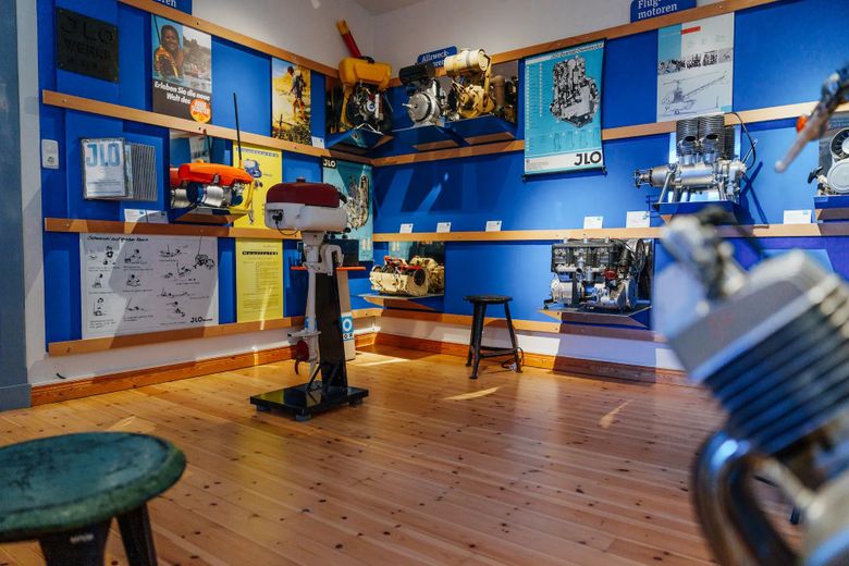 Ein Museumsraum mit blauen Wänden zeigt eine Sammlung von Motoren und technischen Postern. Holzboden und Hocker ergänzen die Ausstellung.