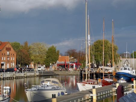 Ein malerischer Hafen mit Segelbooten und historischen Backsteingebäuden. Der Himmel ist bewölkt, und Bäume säumen das Ufer.