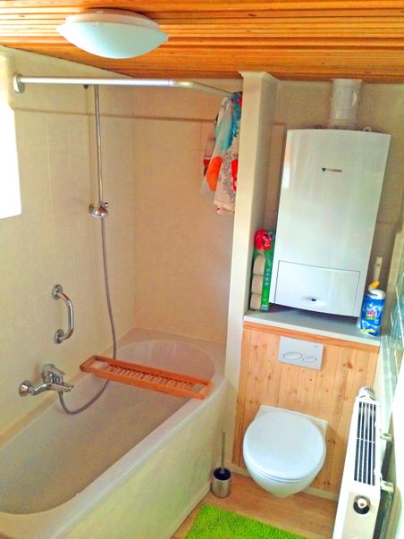 Gemütliches Badezimmer mit Badewanne, Duschvorhang, Toilette und Heizkessel. Helle Holzelemente und grüne Badematte verleihen dem Raum Wärme.