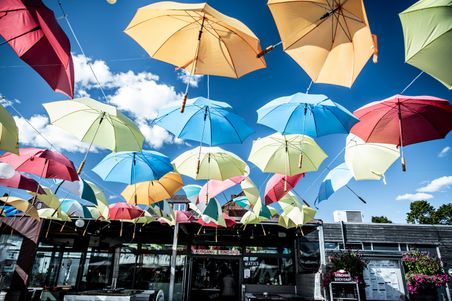 Bunte Regenschirme schweben über einem Café im Freien, aufgehängt an Schnüren. Der Himmel ist blau mit weißen Wolken.