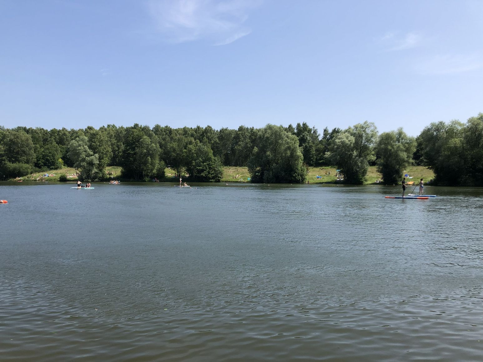 Ein ruhiger See mit Menschen auf Paddelbrettern, umgeben von grünen Bäumen und einem klaren blauen Himmel.