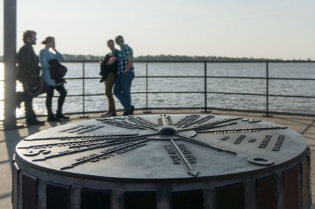 Ein Metallkompass auf einer Mole zeigt verschiedene Richtungen an. Im Hintergrund stehen unscharfe Personen vor einem Gewässer.