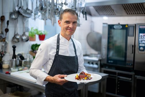 Ein Koch in weißer Uniform und gestreifter Schürze präsentiert ein Gericht in einer professionellen Küche.