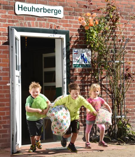 Drei Kinder tragen Kissen aus der Heuherberge vor einer Backsteinwand mit Rosen und offener Tür.