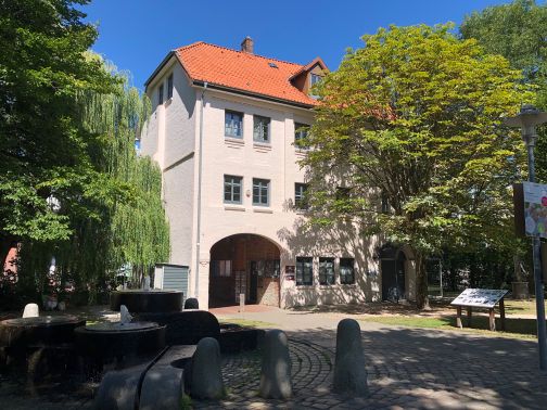 Das Torhaus steht sonnig zwischen Bäumen; im Vordergrund sprudelt ein Brunnen auf dem gepflasterten Platz.
