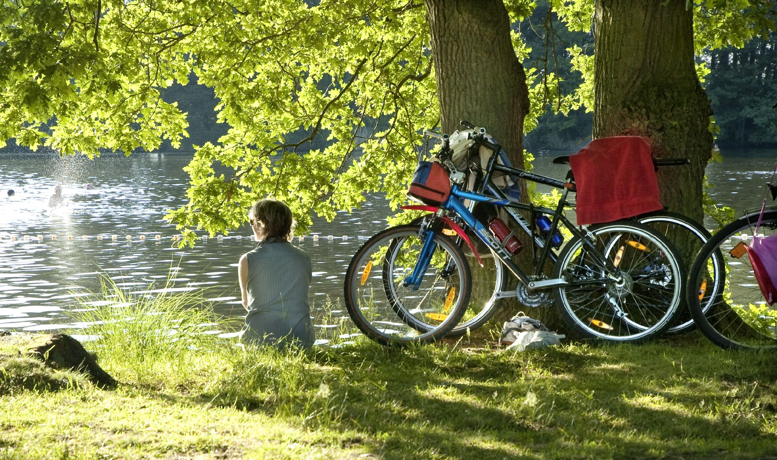 Eine Person sitzt am Ufer eines Sees unter Bäumen, neben zwei Fahrrädern. Im Hintergrund schwimmen Menschen im Wasser.