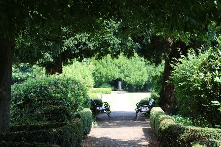 Ein schattiger Gartenweg mit zwei Bänken, umgeben von üppigem Grün und Bäumen. Im Hintergrund ist ein Denkmal zu sehen.