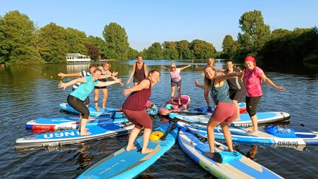 Eine fröhliche Gruppe von Menschen steht auf Stand-Up-Paddle-Boards auf einem See. Sie posieren lachend in der Abendsonne, umgeben von grünen Bäumen.
