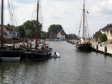 Historische Segelboote liegen im ruhigen Binnenhafen vor Anker, umgeben von traditionellen Gebäuden und grünen Bäumen.
