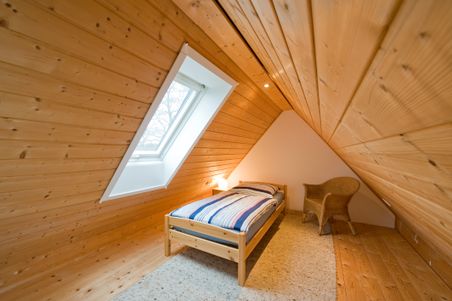 Gemütliches Dachzimmer mit Holzwänden, einem Einzelbett, einem Korbstuhl und einem Dachfenster, das Tageslicht hereinlässt.
