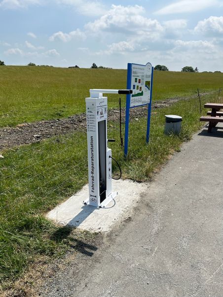 Fahrrad-Reparaturstation am Radweg neben Infotafel, Wiese und Picknickbank unter blauem Himmel.