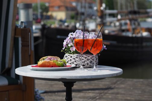Ein Matjesbrötchen und zwei Aperol Spritz auf einem Tisch am Hafen, dekoriert mit einem Blumenkorb.